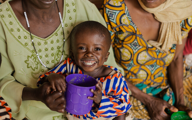 Der Junge lächelt nach seiner Genesung im Ernährungszentrum © Fairpicture Ein kleiner Junge mit einem Becher Brei lächelt auf dem Schoß seiner Mutter – er wurde im Ernährungszentrum Gitega aufgepäppelt