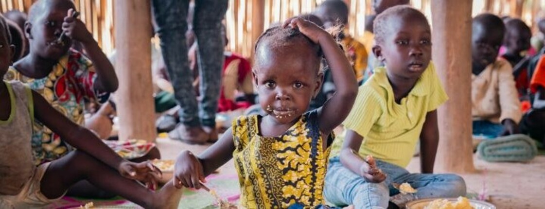 Kinder sitzen auf einer bunten Matte und essen gemeinsam in einer Hilfseinrichtung. Im Hintergrund sind weitere Kinder zu sehen.